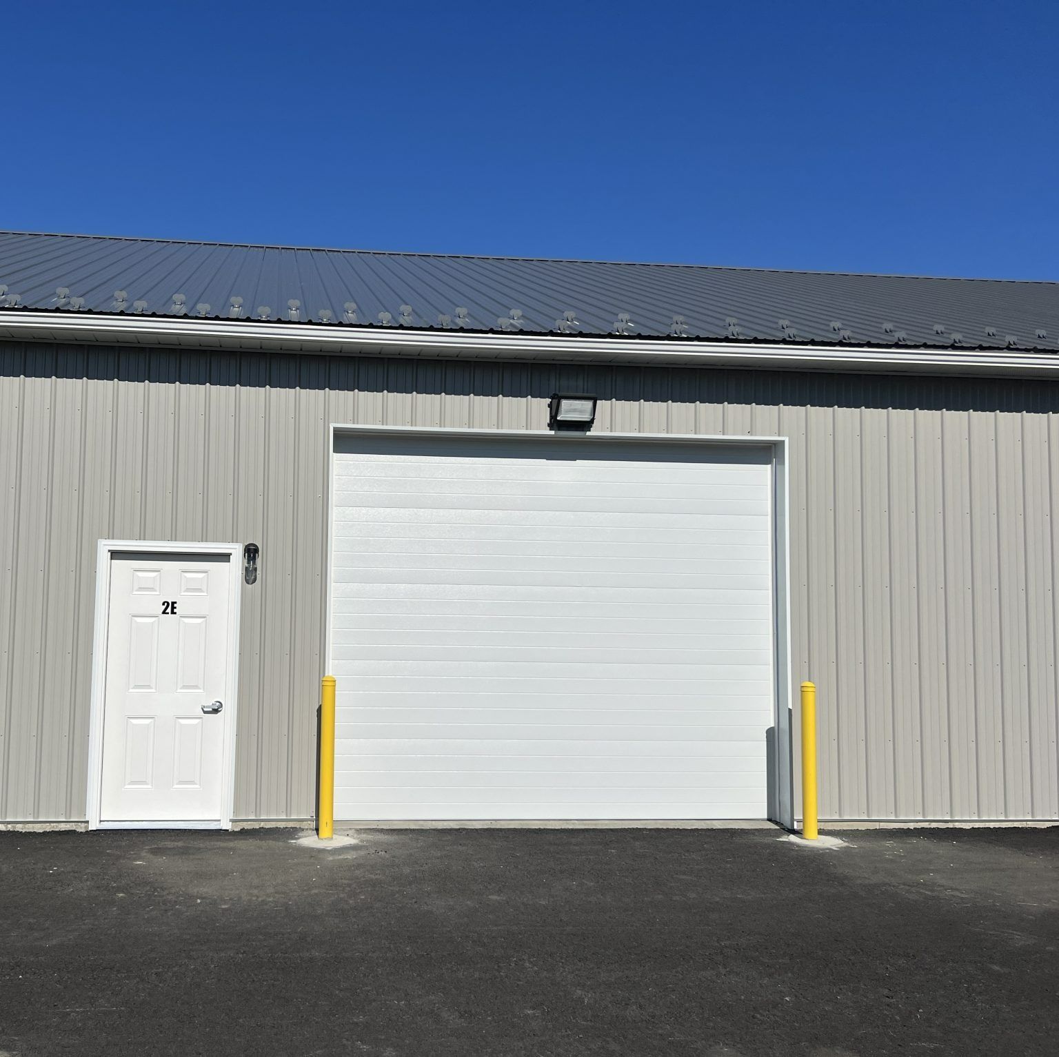 Gray industrial building with a white garage door, side entrance, and two yellow bollards under a blue sky