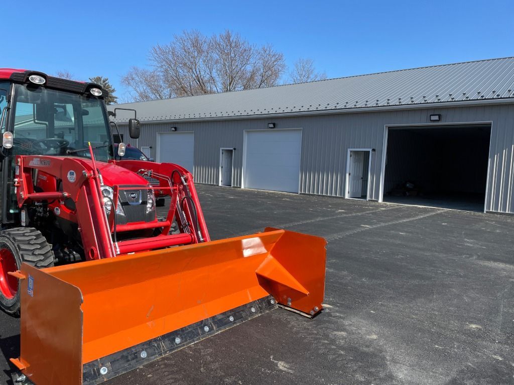 Red tractor with orange snowplow parked outside a gray warehouse on a sunny day