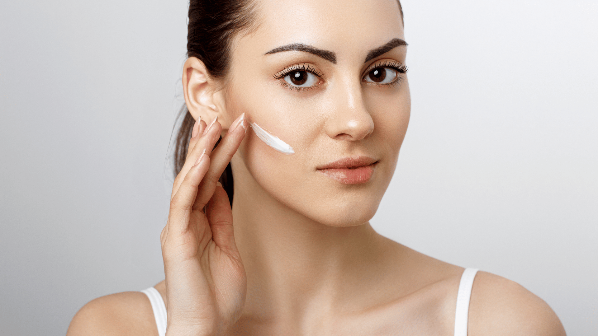 Woman applying white cream to her cheek with a clean, neutral expression