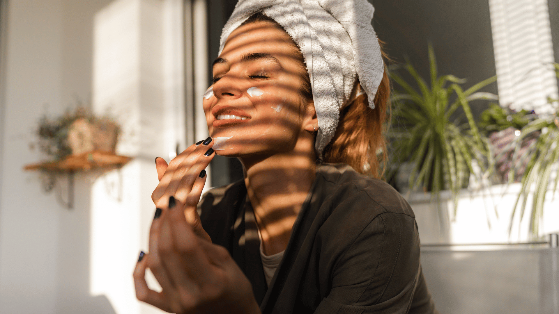 Woman smiling by a sunlit window, wearing a towel and black top, with plant shadows across her face.