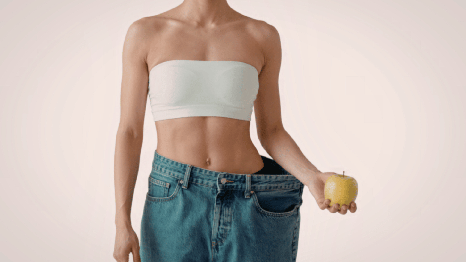 Person in a white tube top and loose jeans holding a green apple against a plain background
