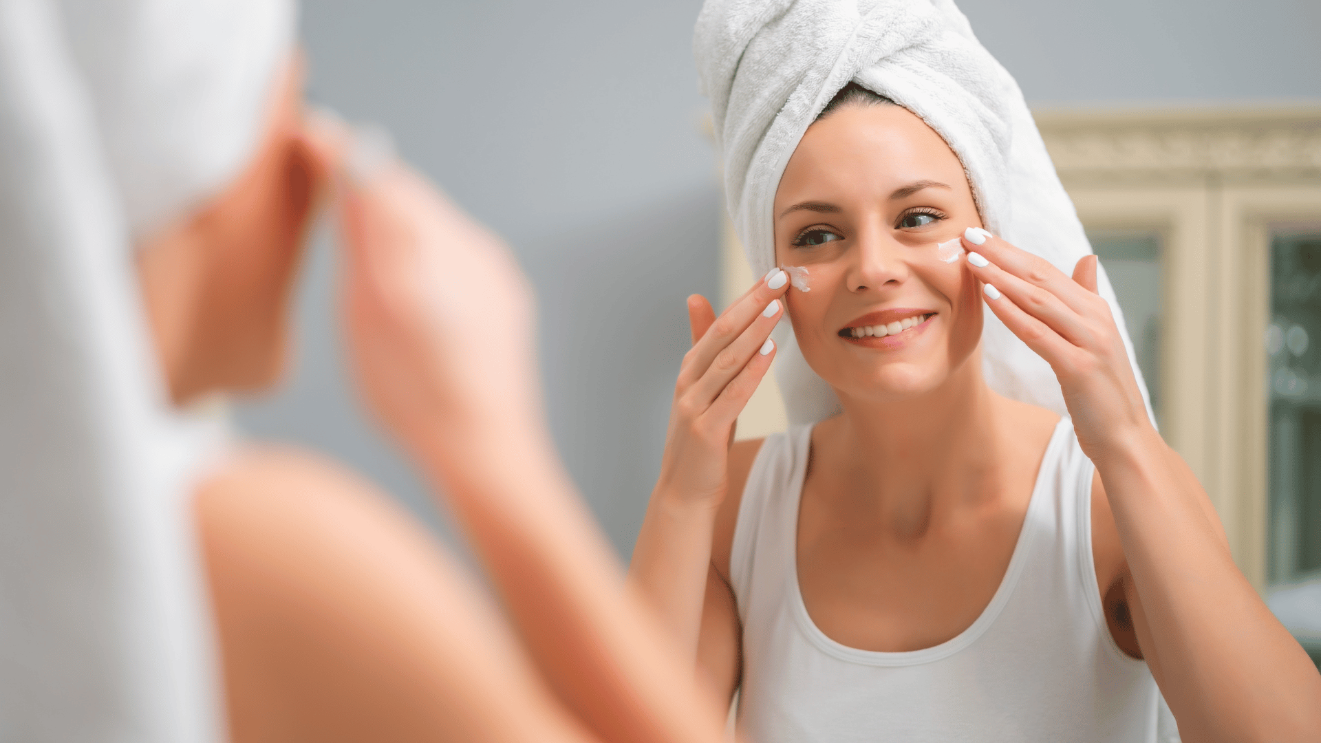 Person applying face cream in a bathroom mirror, with a white towel wrapped around their head.