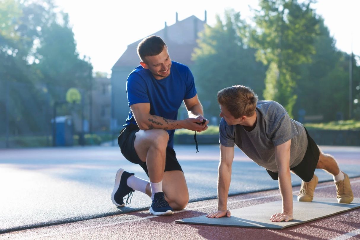 Two people stretching on a mat outdoors, with one kneeling and the other in a plank position.