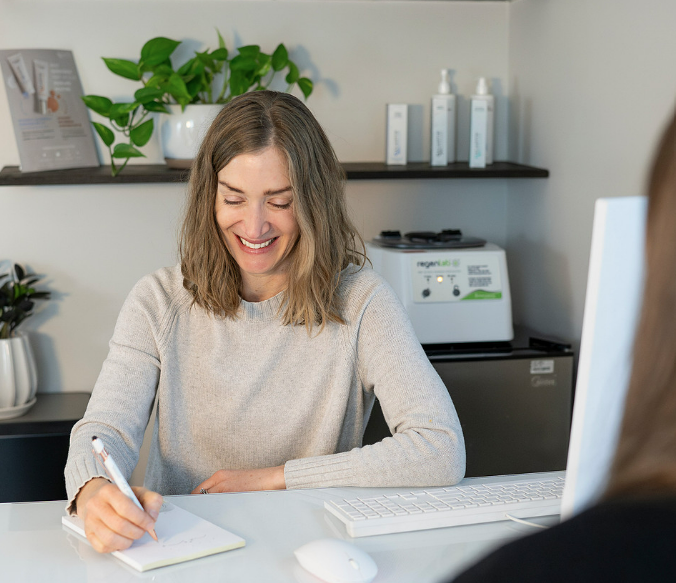 Woman smiling, writing at a desk, looking down. Office setting with plants and equipment.