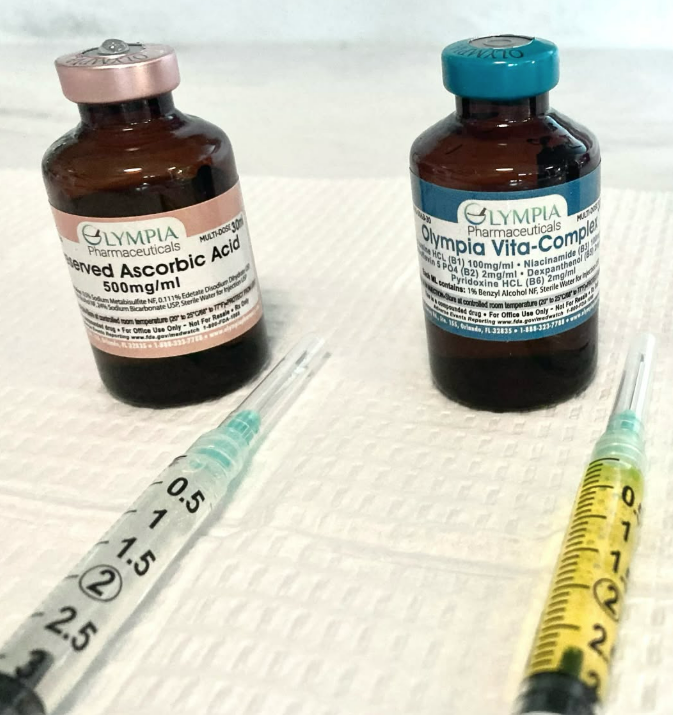 Two vitamin C vials with syringes on a white surface, one brown bottle and one clear syringe with yellow liquid.