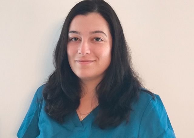 Woman with dark hair in a teal top smiles, facing the camera against a light background.