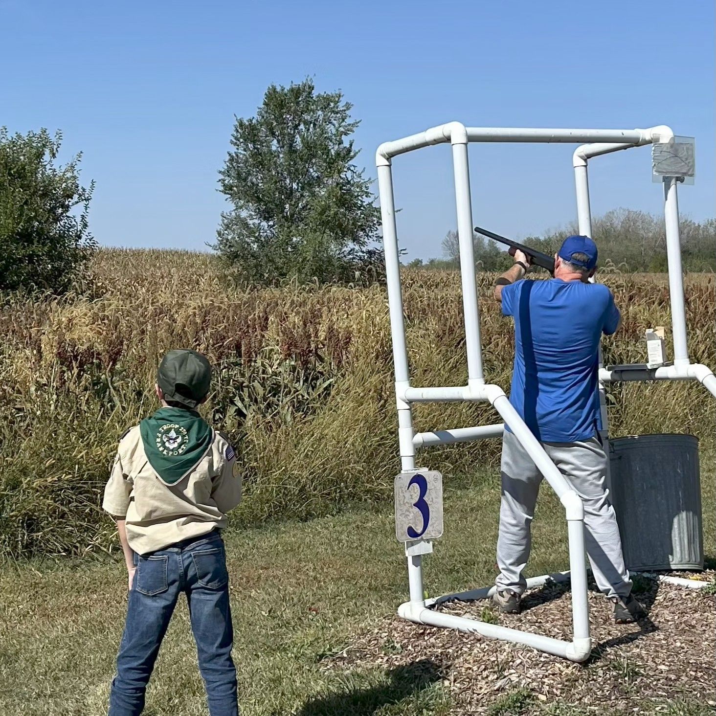 Man shooting a shotgun at a skeet range, overseen by a boy scout, outdoors. Sunny day.