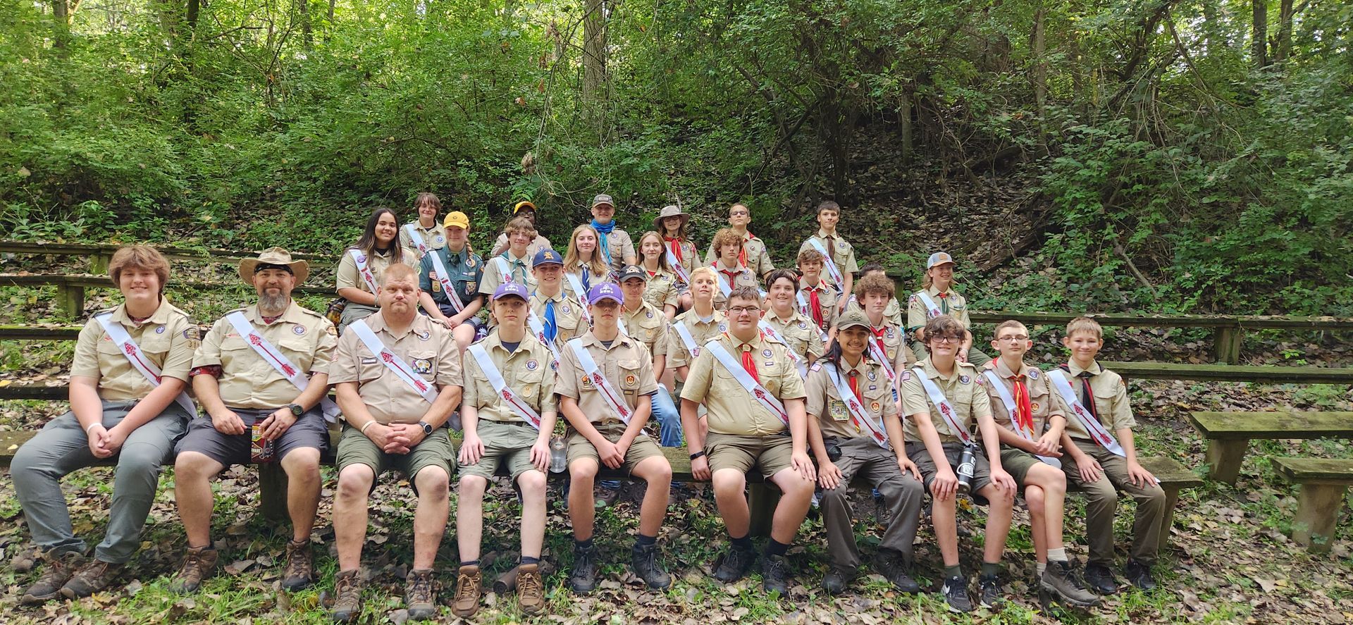 A group of boy scouts are sitting on a bench in the woods.