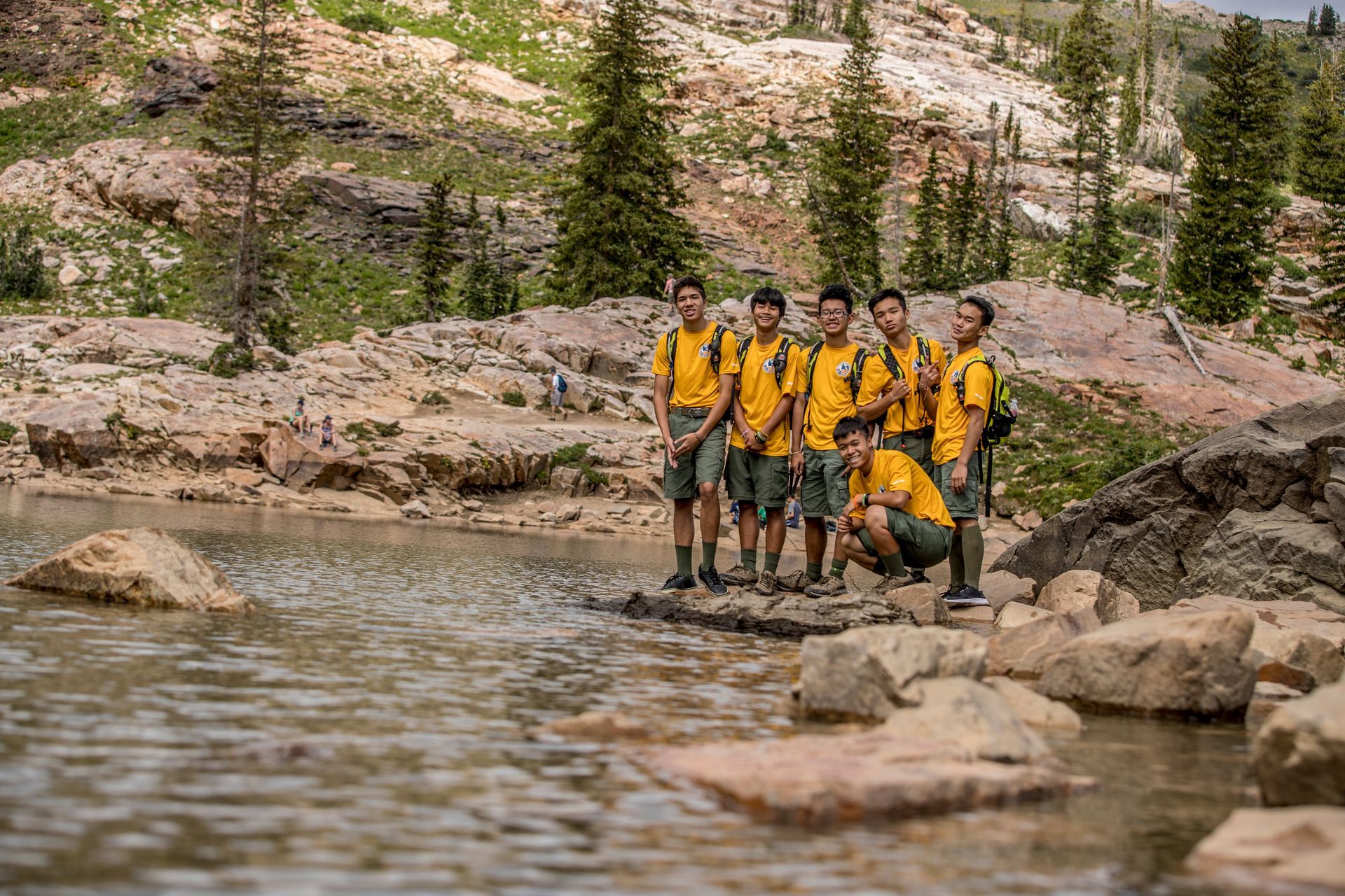 Group of six people in yellow shirts and green shorts near a lake, rocky terrain, trees, and mountain.