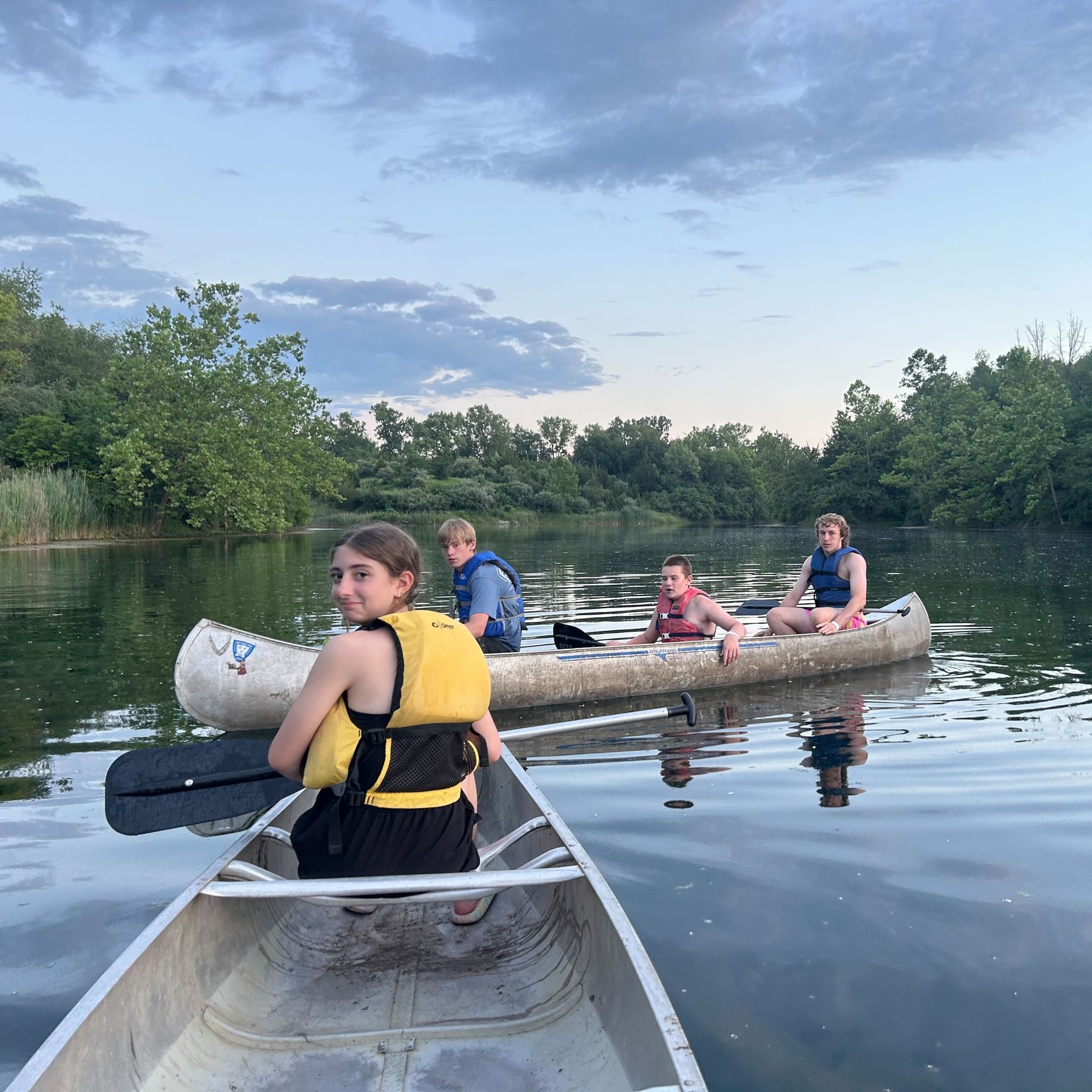 Four people canoeing on calm water; two canoes. Sky is overcast with trees on the banks.