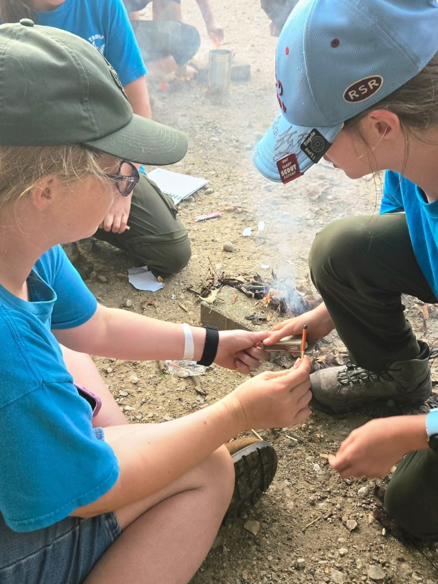Two girls in blue shirts and caps, lighting a small fire with matches outside.