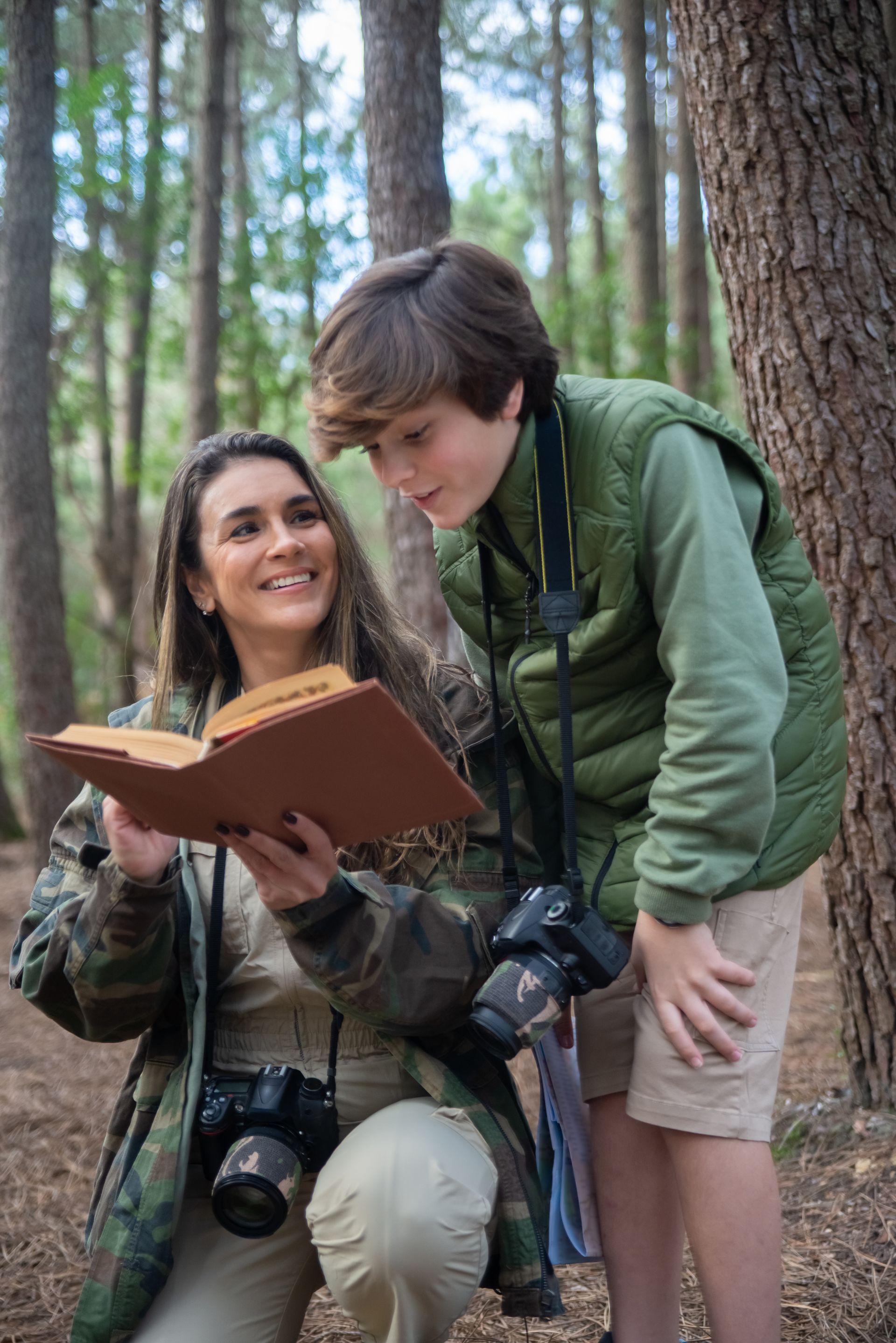 Woman and boy in forest looking at open book; both wear cameras.
