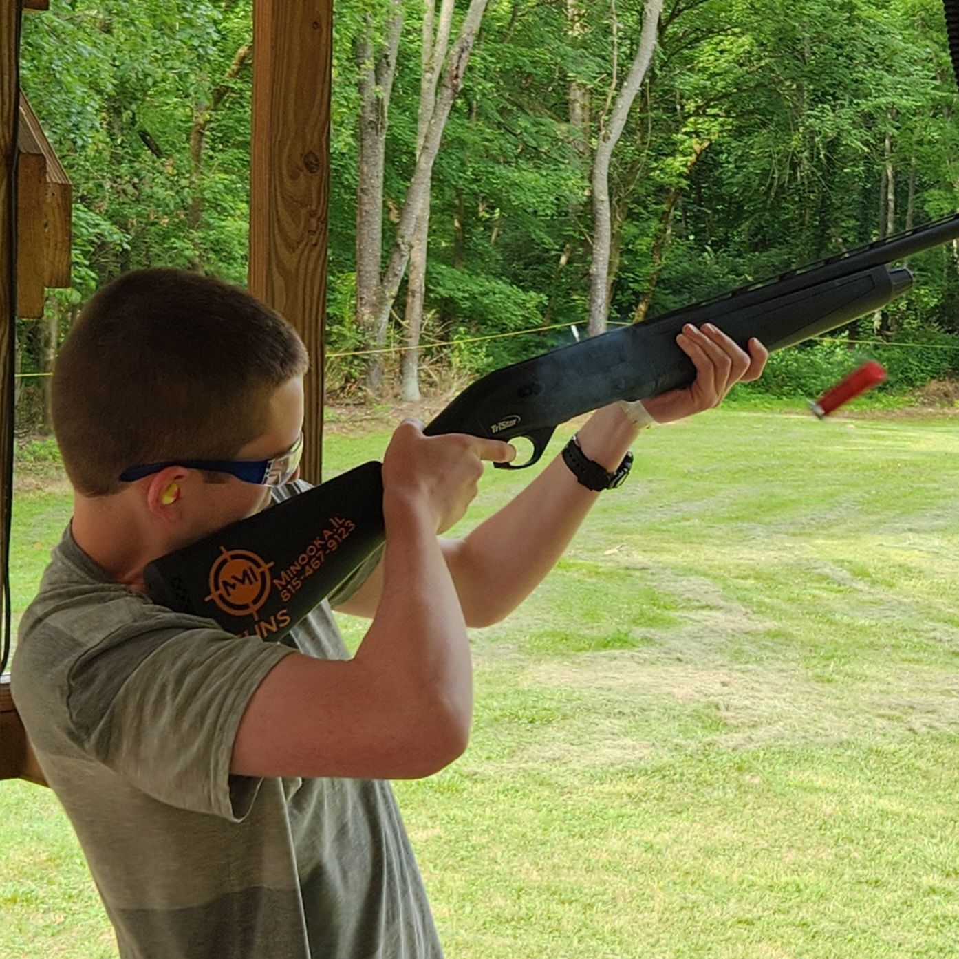 Person aiming a shotgun, wearing safety glasses and ear plugs, outdoors.
