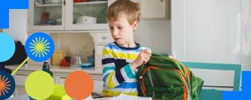 Child packing a green backpack at a table with colorful classroom supplies