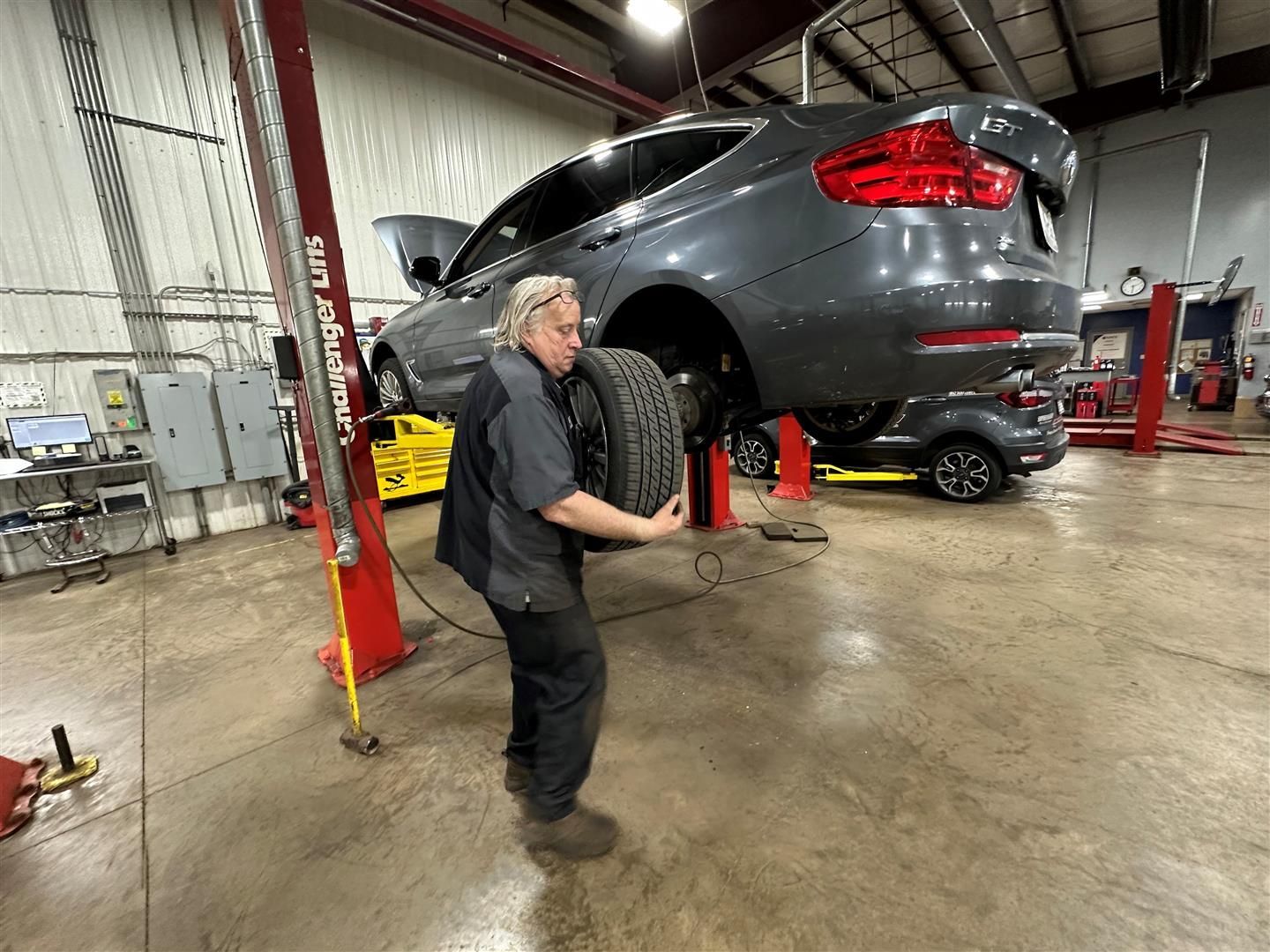A man is holding a tire in front of a car on a lift in a garage. | Lou's Car Care Center, Inc.