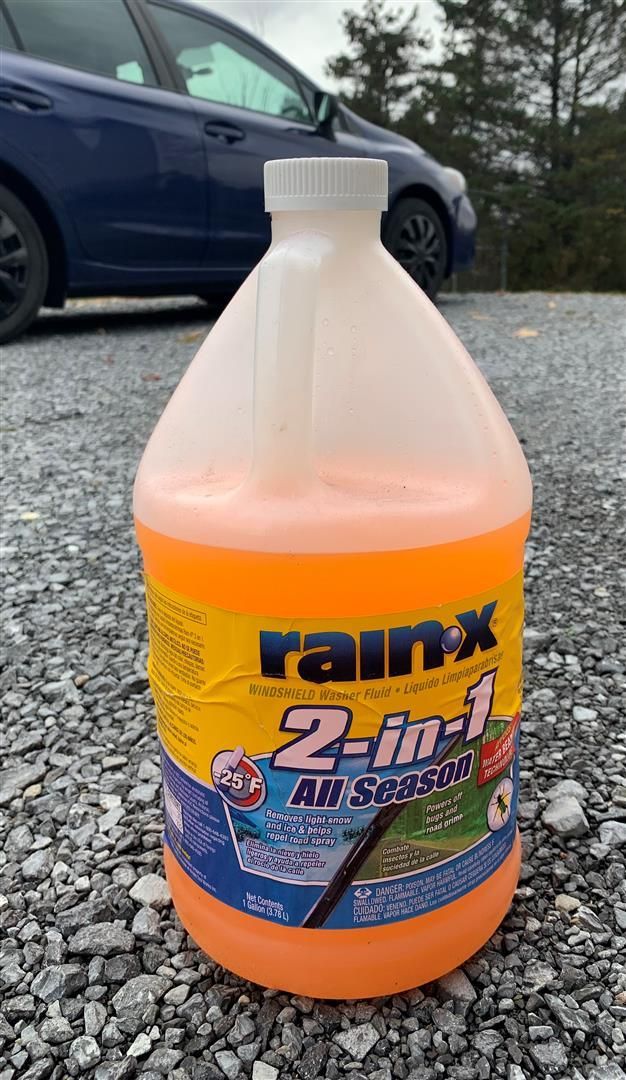 A gallon of rain x 2 in 1 all season sitting on gravel in front of a blue car. | Lou's Car Care Center Inc.