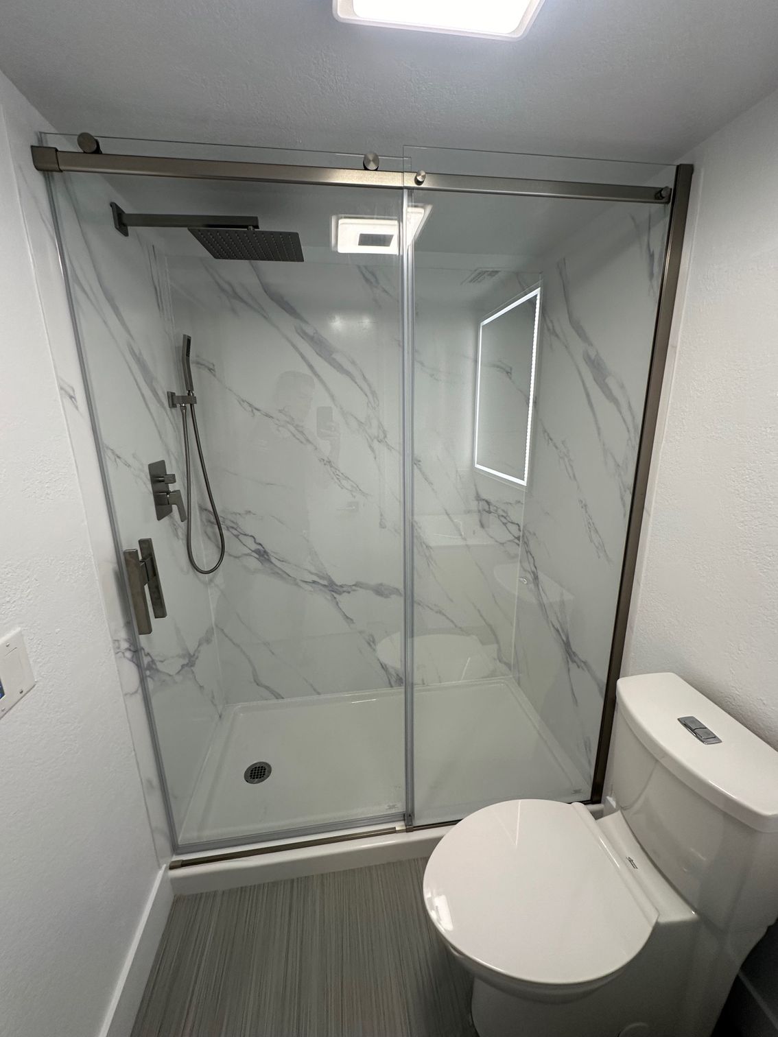 Bathroom with marble-look shower and glass door, next to a white toilet on a gray floor.