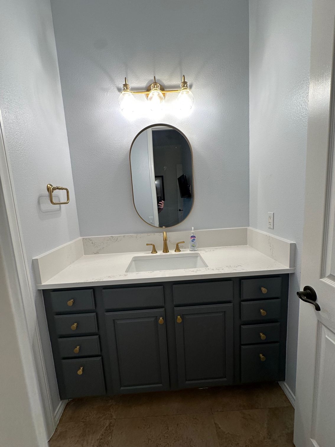 Bathroom with gray vanity, gold fixtures, oval mirror, and light blue walls.
