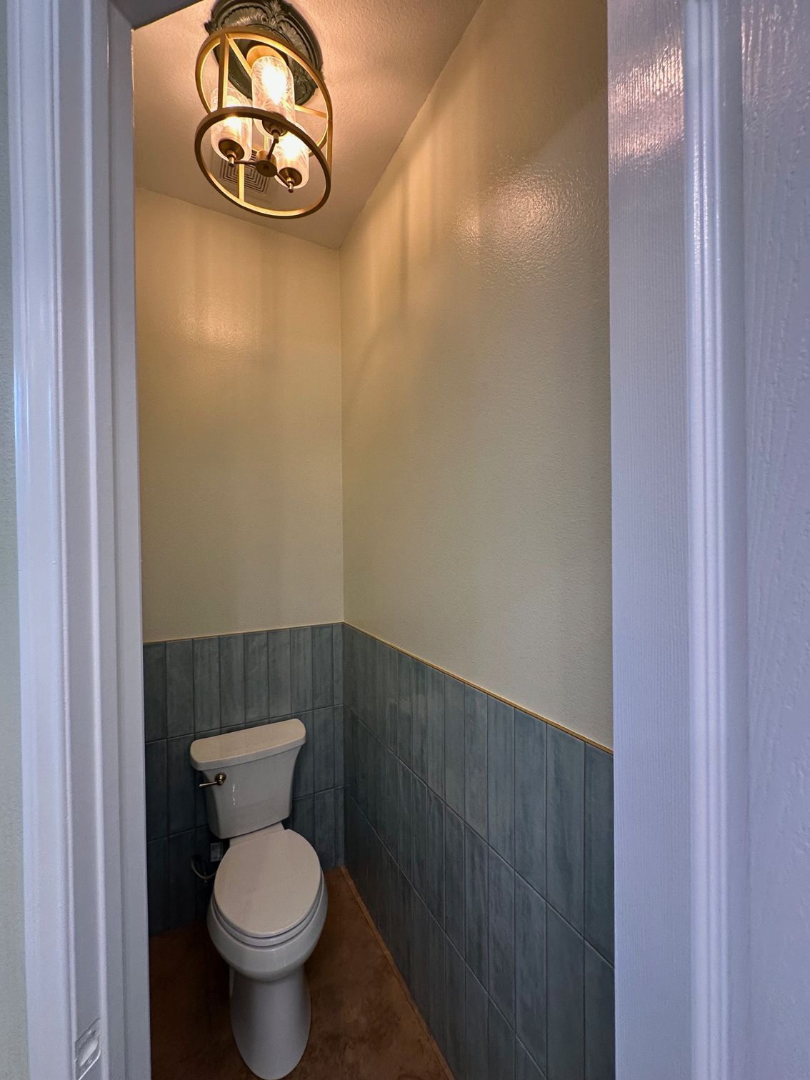 Small bathroom with a toilet and blue tiled wainscoting, under a gold-colored light fixture.