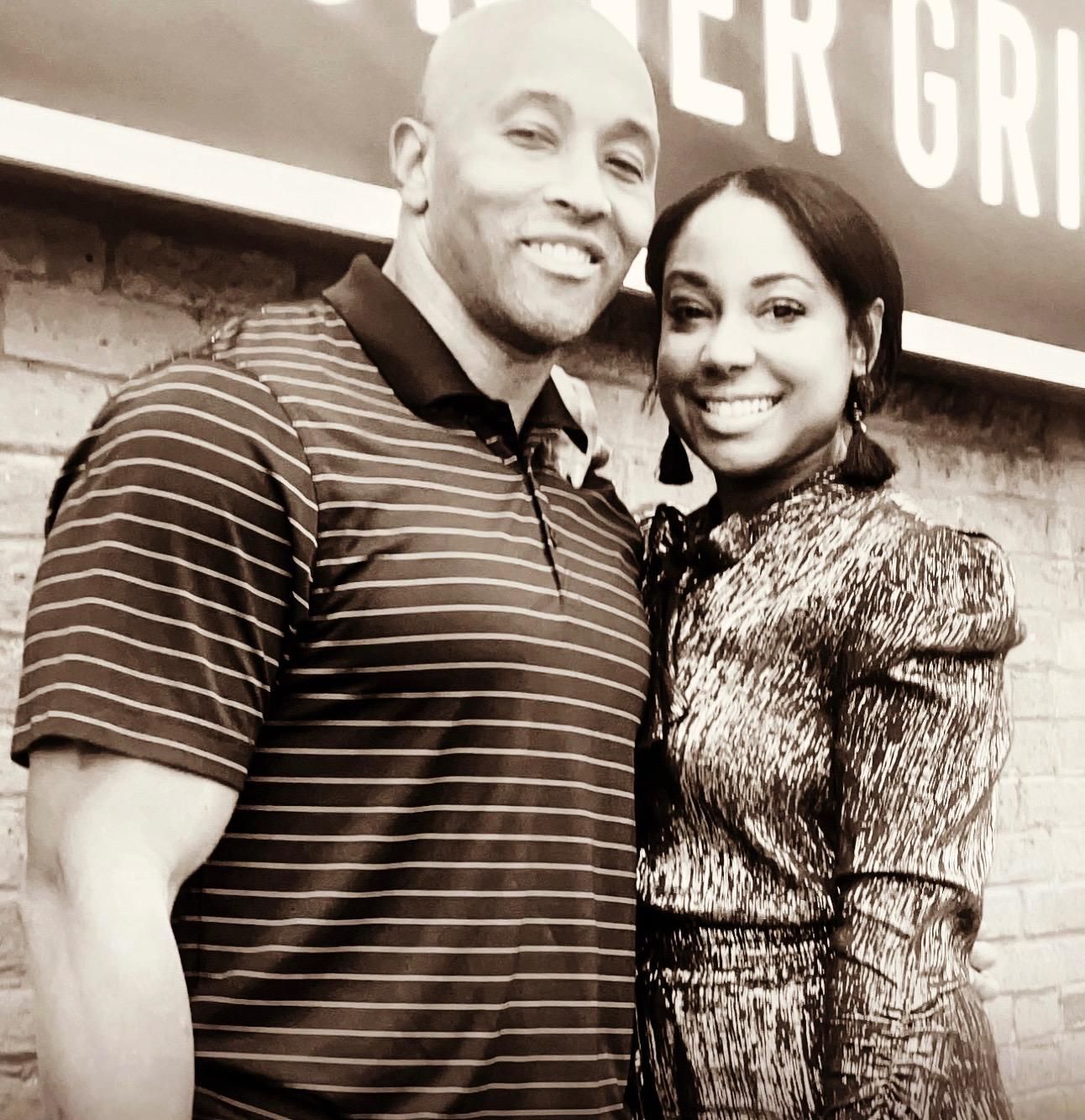 Man and woman smiling, posed closely together in front of a brick wall, possibly at a restaurant.