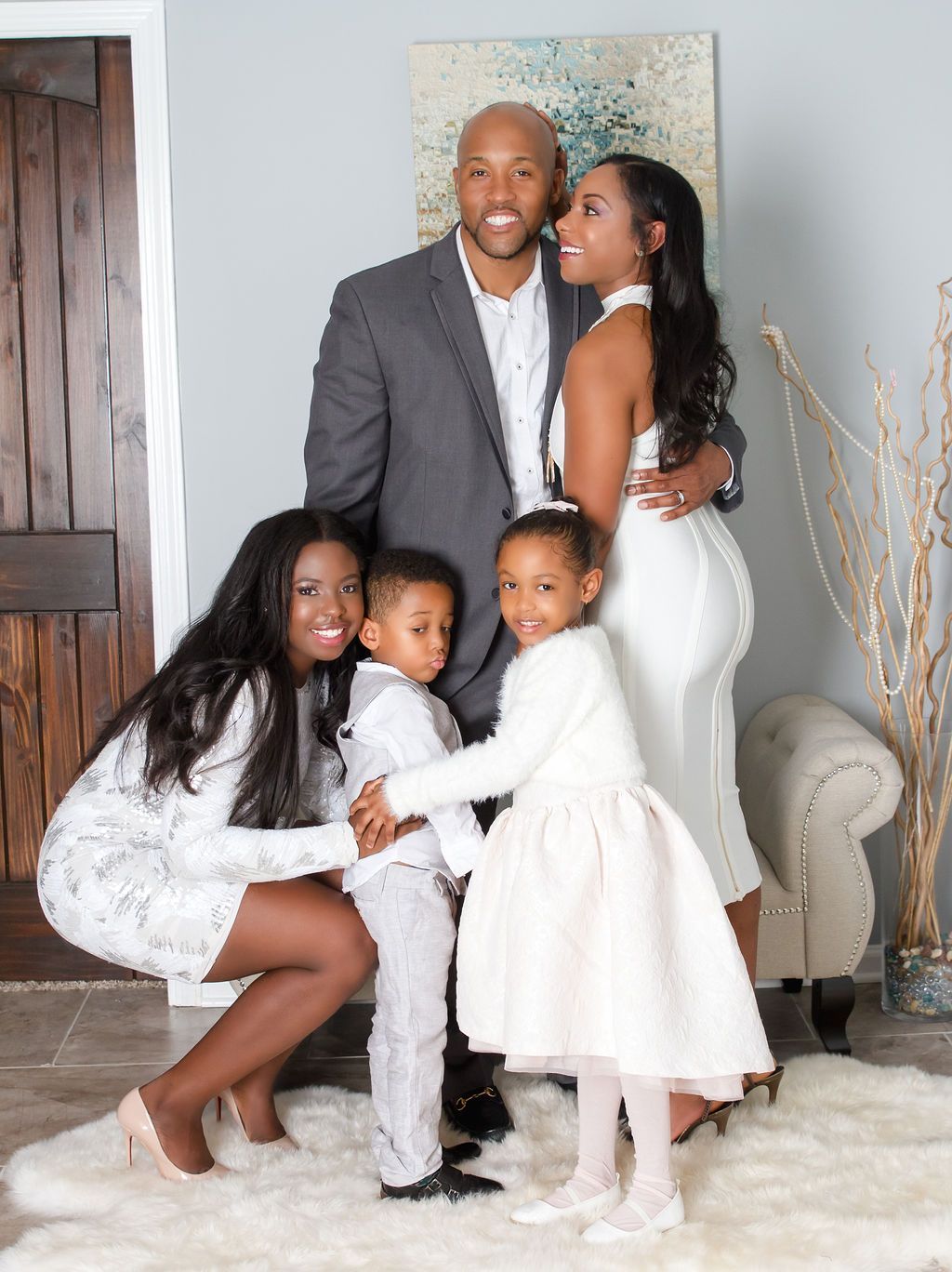 Family of five poses indoors: woman kneeling, two small children, and couple embracing.