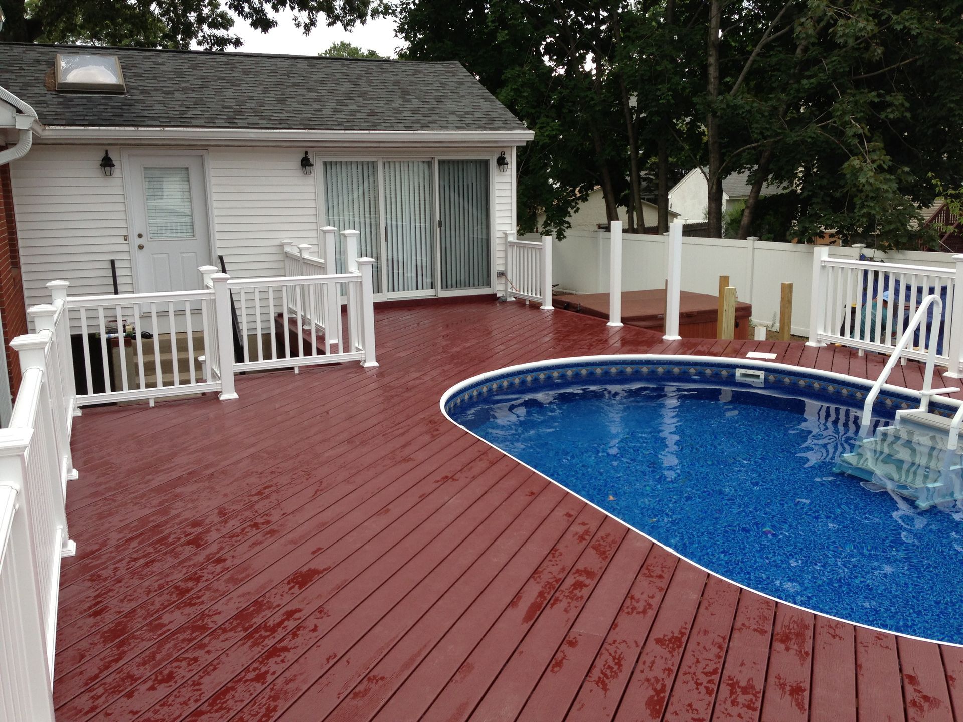 Red wooden deck with white railing surrounding an oval pool. House with sliding glass door.