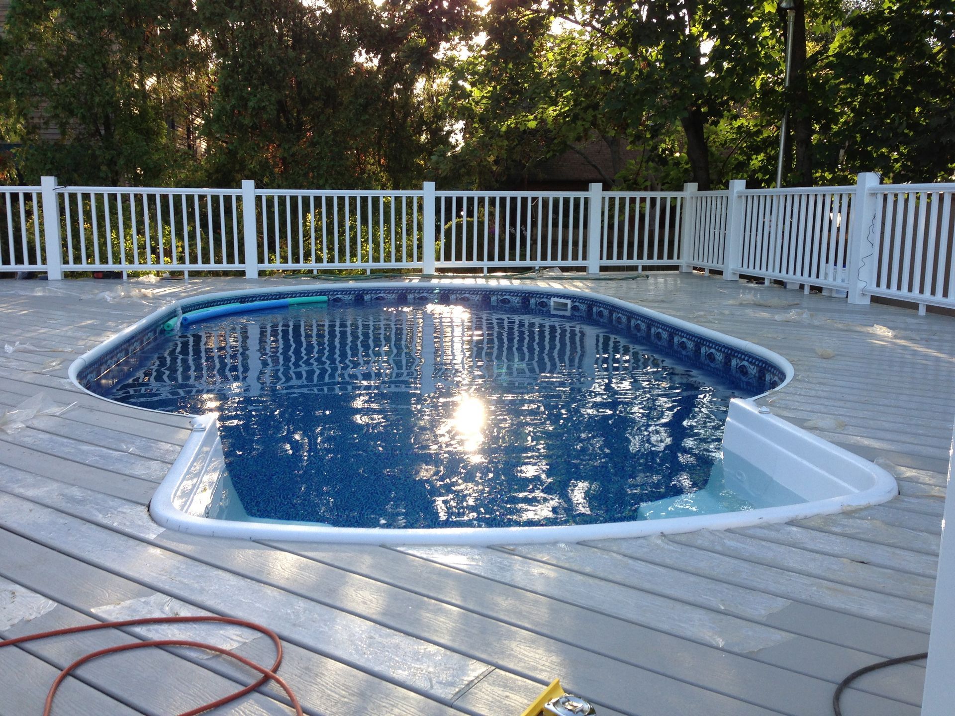 Above-ground swimming pool on a wooden deck, surrounded by a white fence. Sunlight reflects on the water.