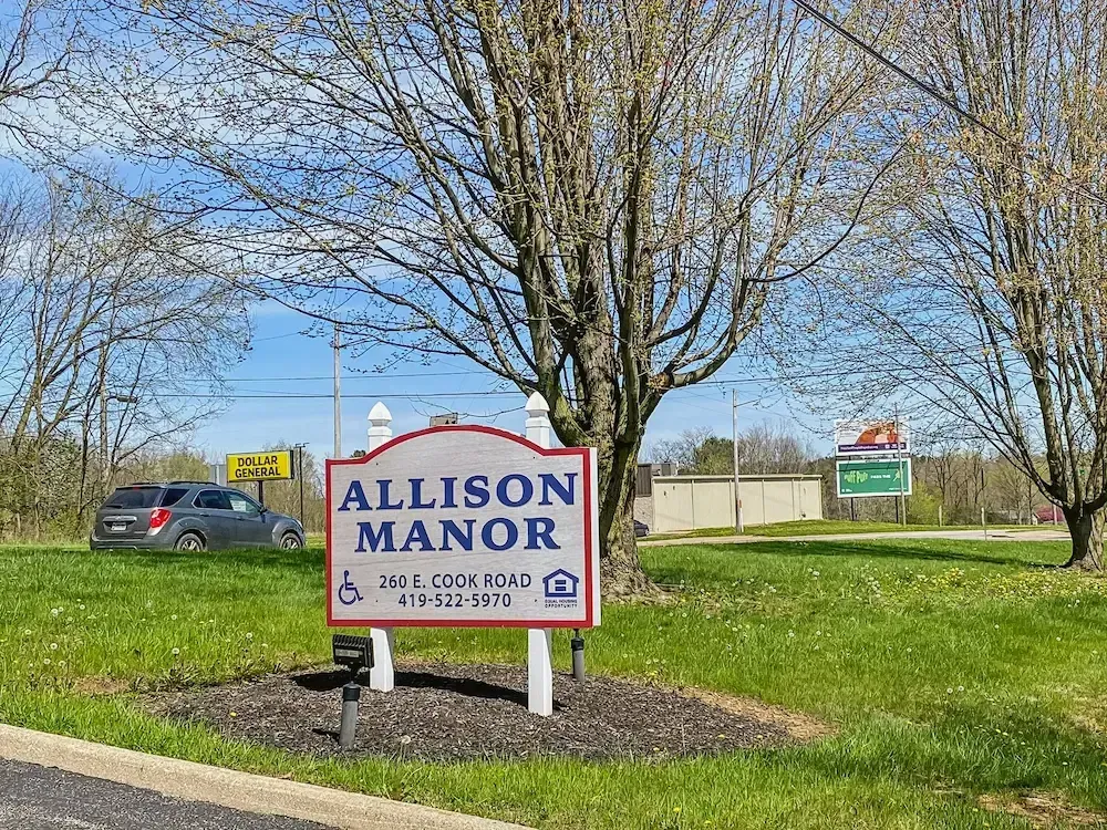 A sign for allison manor is sitting in the middle of a grassy field.