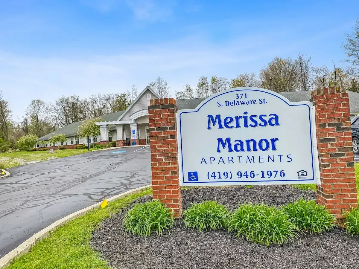 A sign for merissa manor apartments is in front of a brick building.