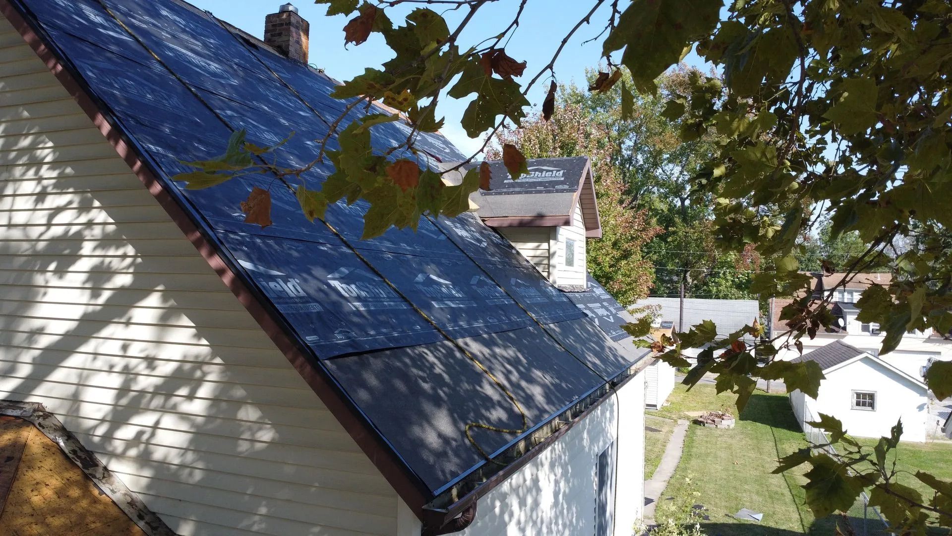Solar panels on a sloped roof. Sunlight casts shadows on the panels and the white house. Autumn leaves frame the view.