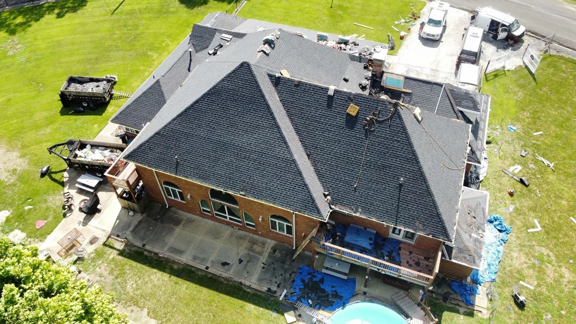 Aerial view of a house with roof damage, surrounded by debris. Blue tarp covers the pool.