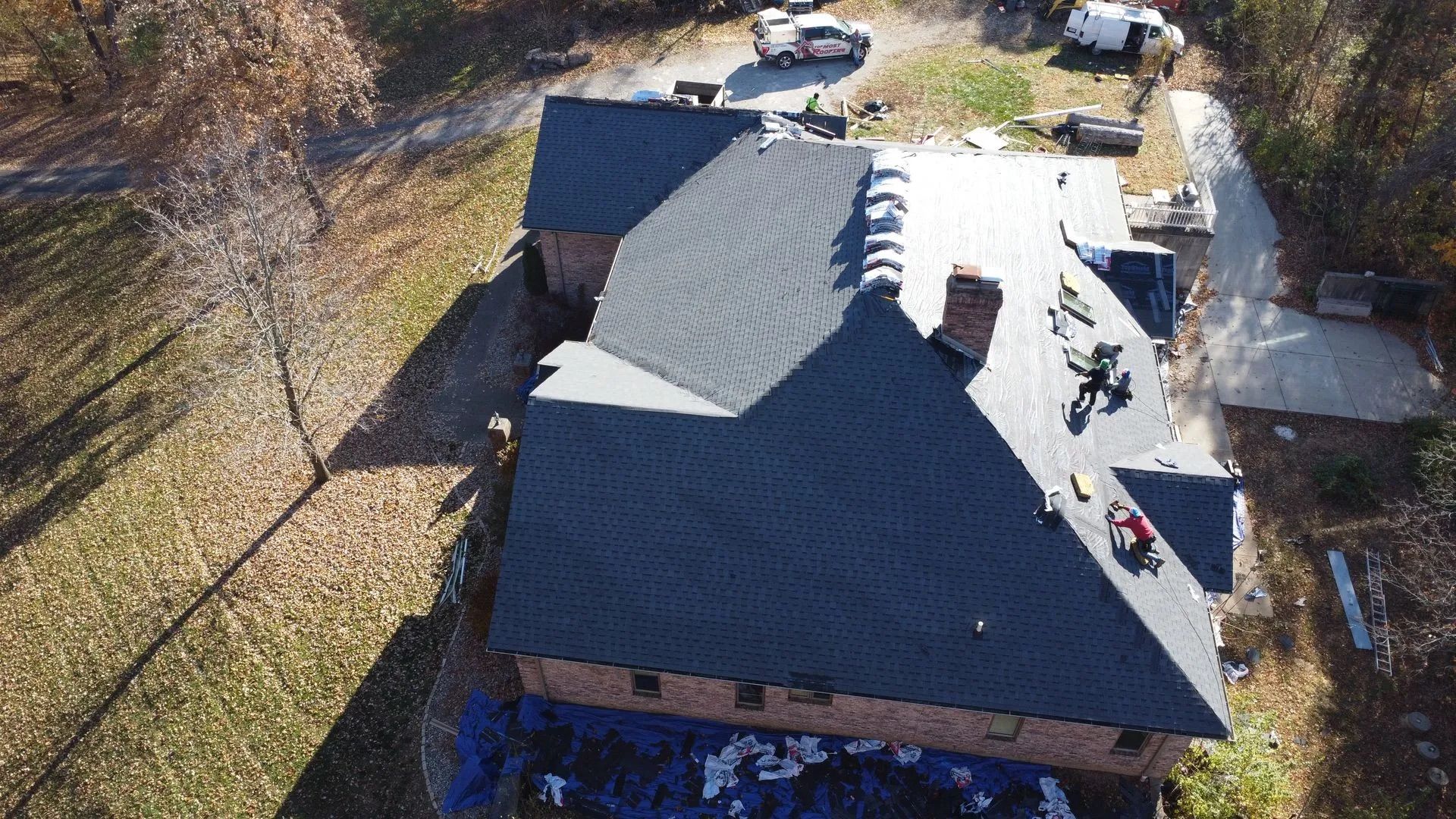 Aerial view of a house roof partially covered with dark shingles, workers visible, setting is outdoors.