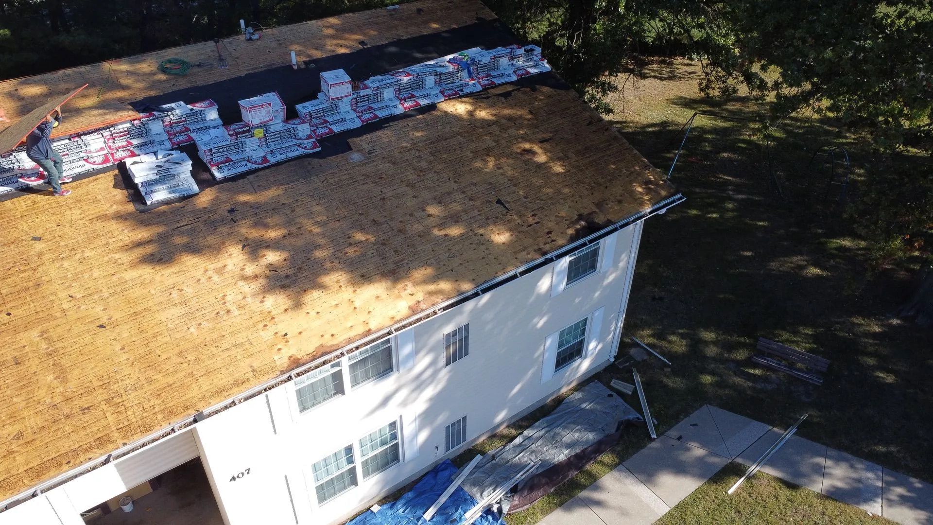 Roofing work in progress on a two-story house. Shingles and supplies on the roof, sunlight casting shadows.
