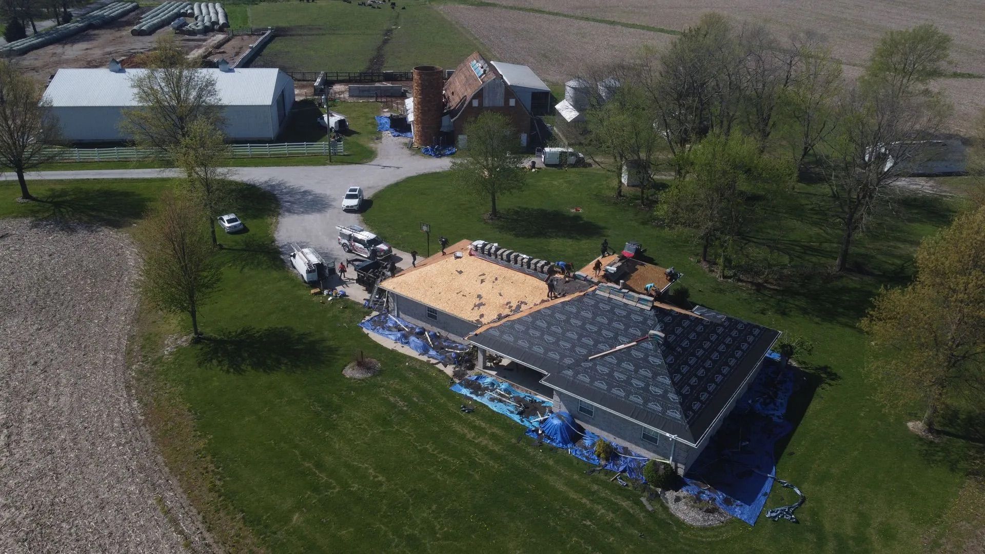 Aerial view of a house with roofing being done; blue tarps, green lawn, farm buildings in background.