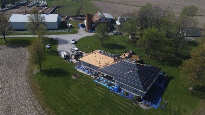 Aerial view of a home with a roof under construction on a grassy property. A farm is in the background.