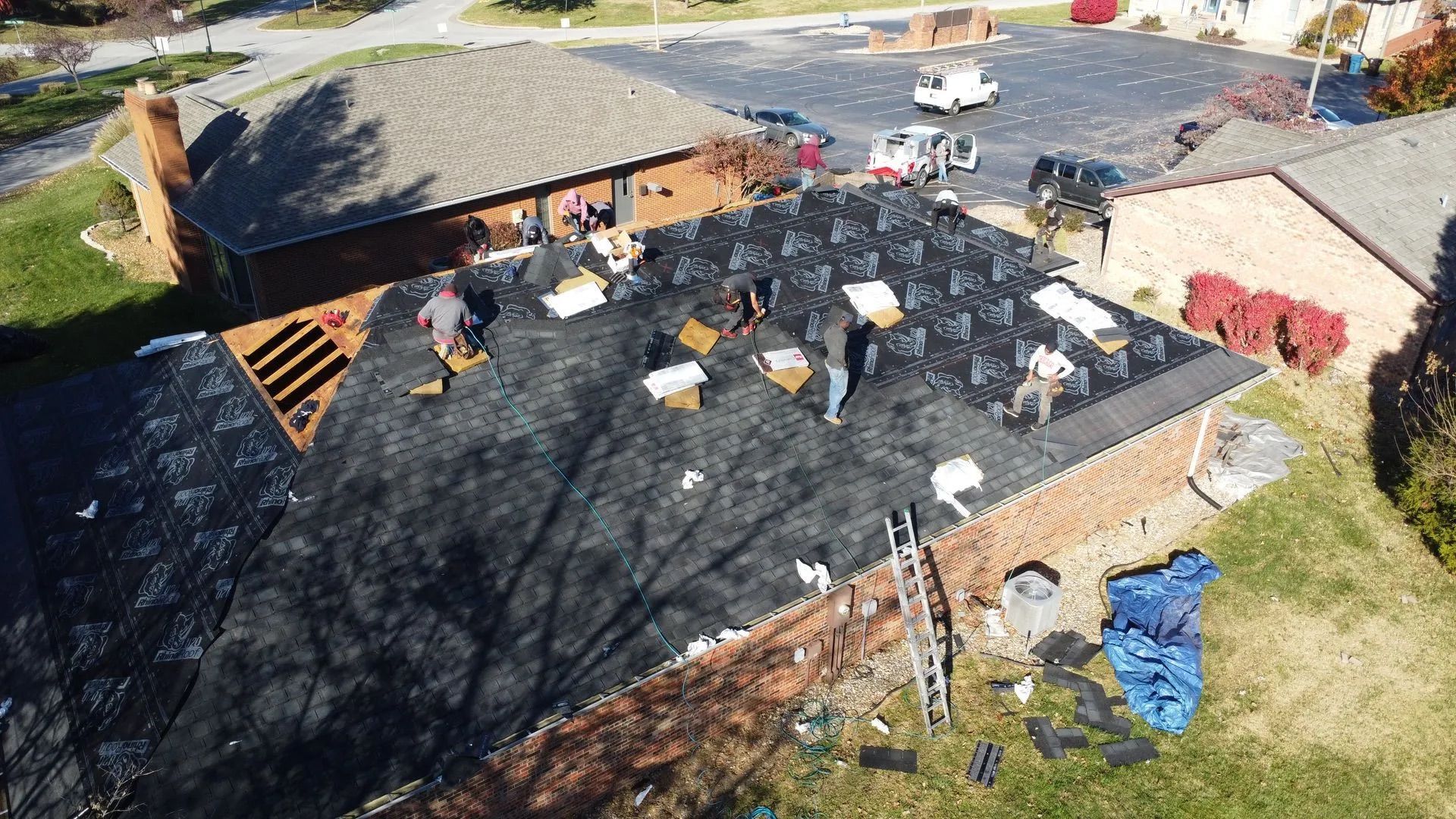 Roofing crew working on a dark shingle roof of a building with surrounding brick and other buildings.