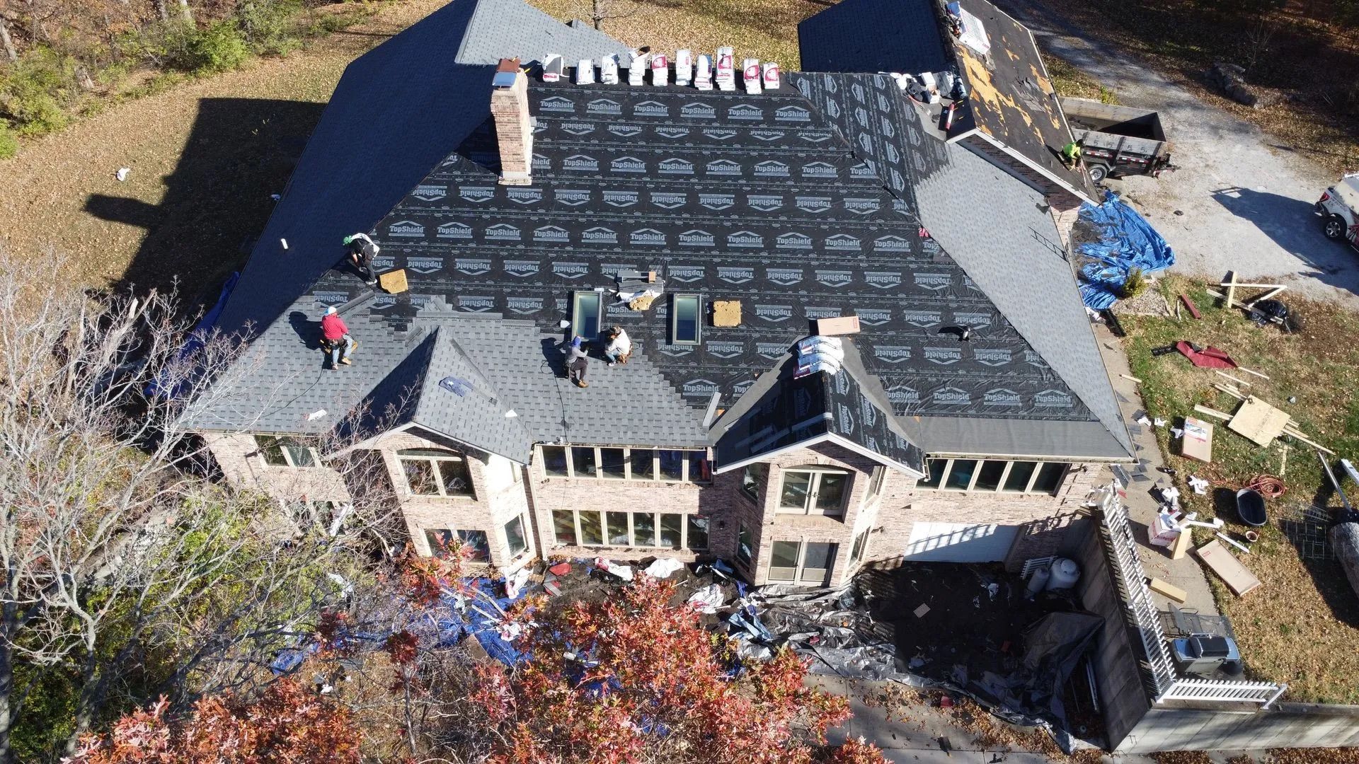 Workers on a large home's roof, replacing shingles. Brown brick exterior, trees surround the house.