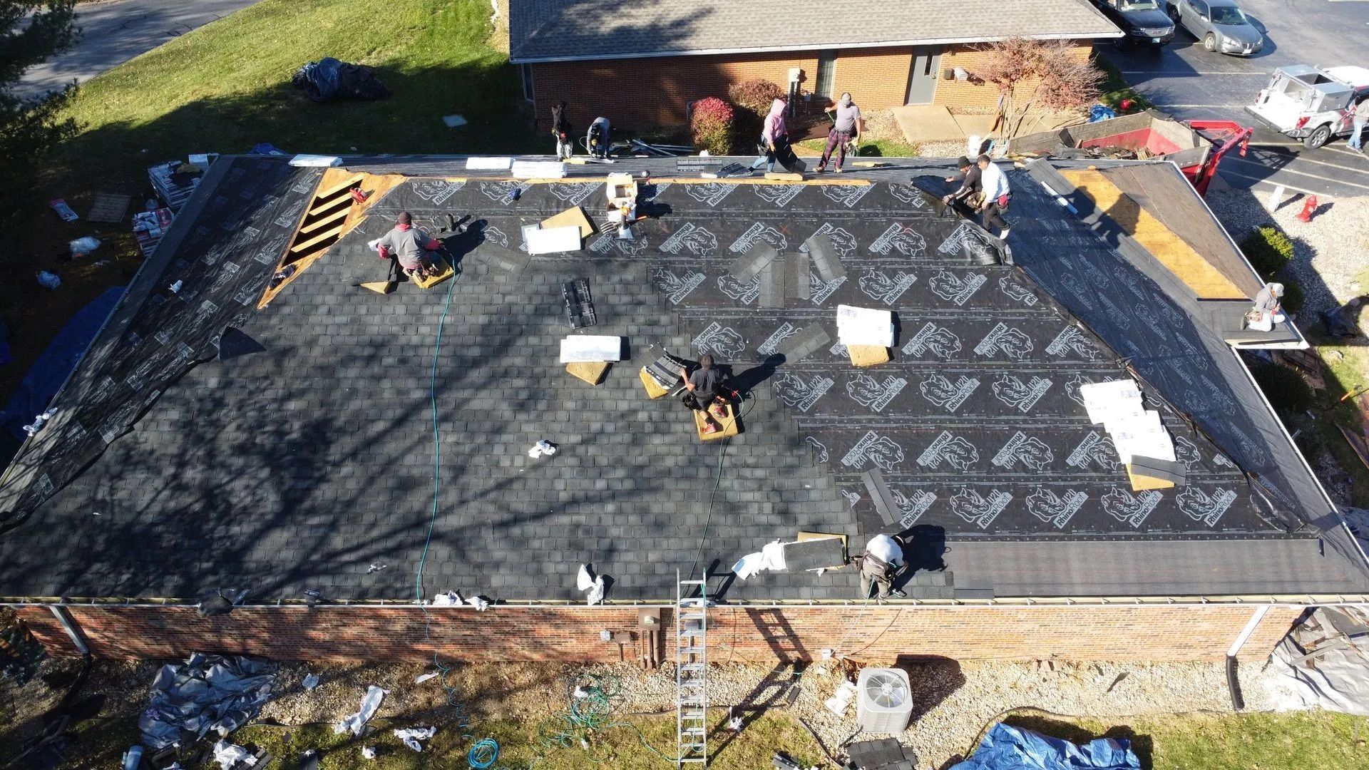 Roofers working on a flat roof, applying dark material. Other buildings and green surroundings visible.