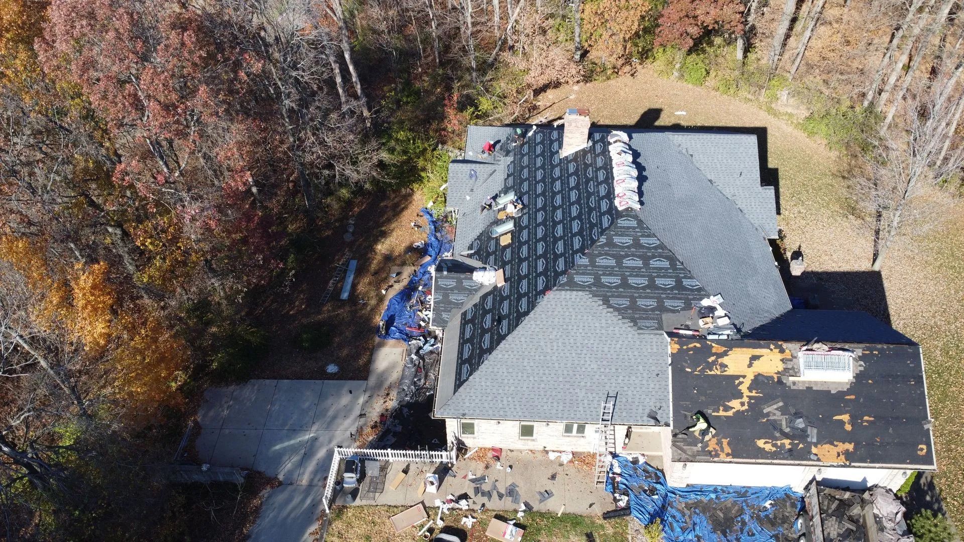 Aerial view of a house with roofing in progress. Blue tarps cover the yard and roof. Fall foliage surrounds it.