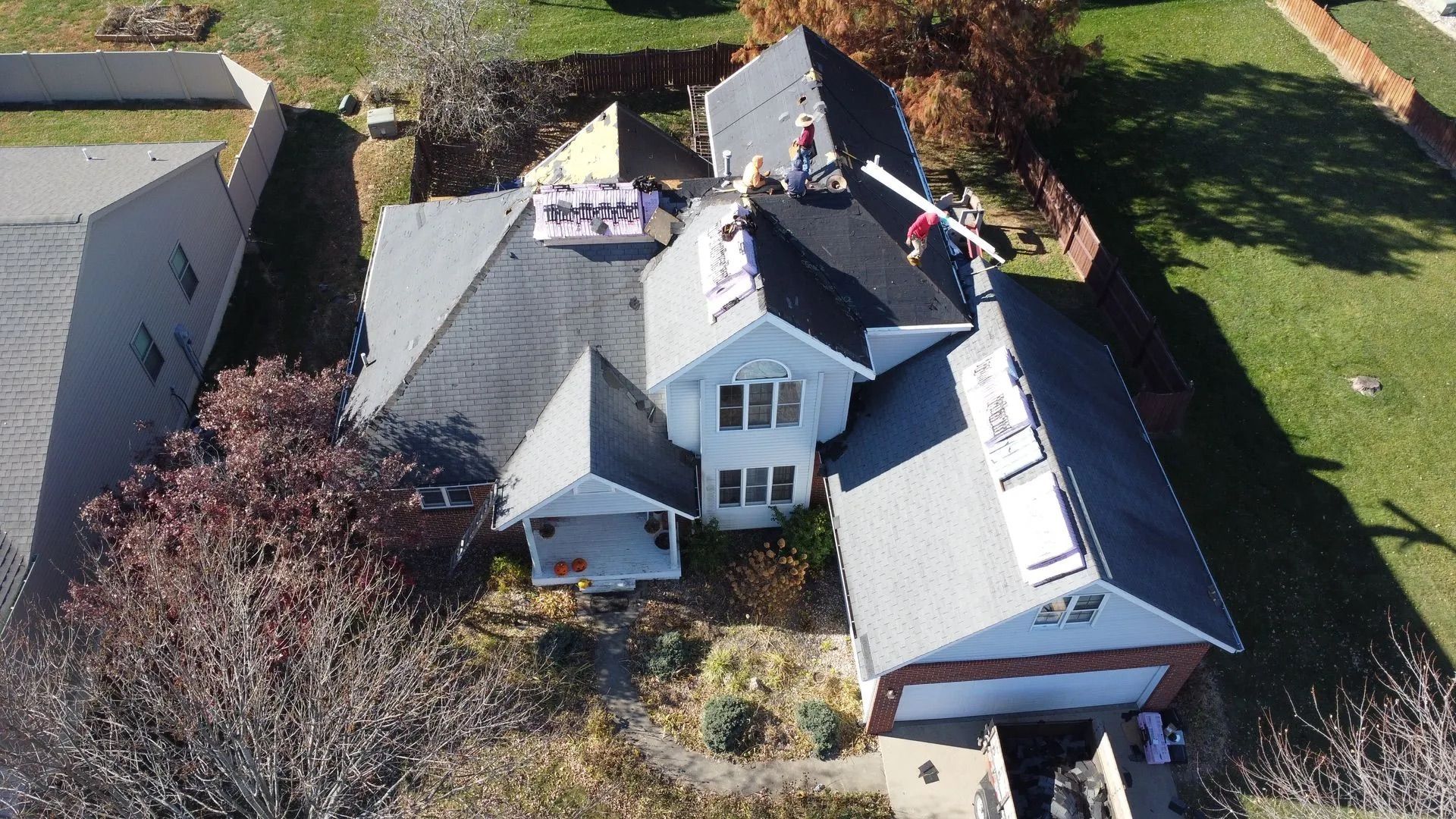 Workers on a roof installing shingles; house with gray roof, brick garage, and fall foliage.