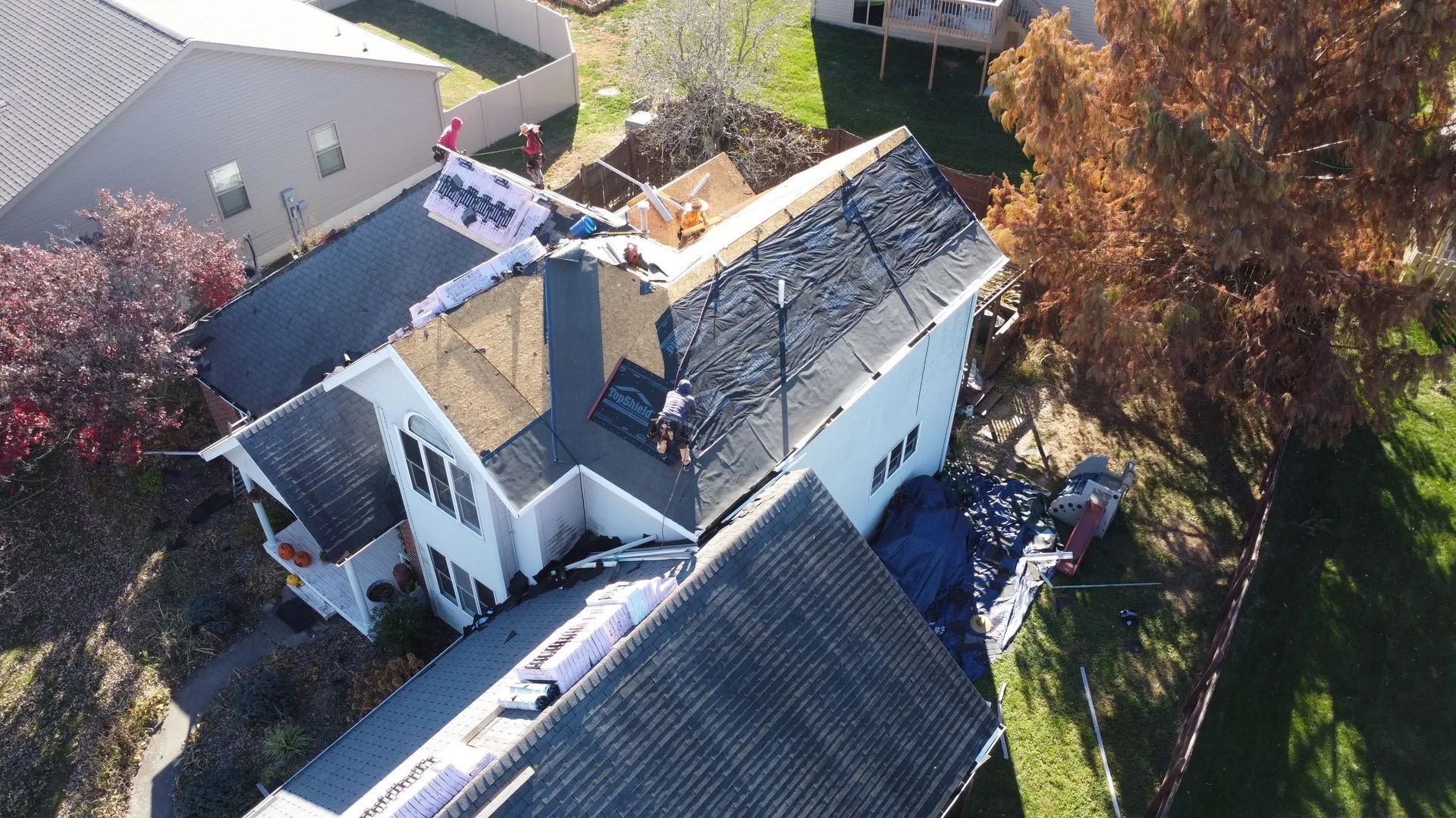 Aerial view of house with roofing being replaced; partially covered with blue tarp, surrounding yard.