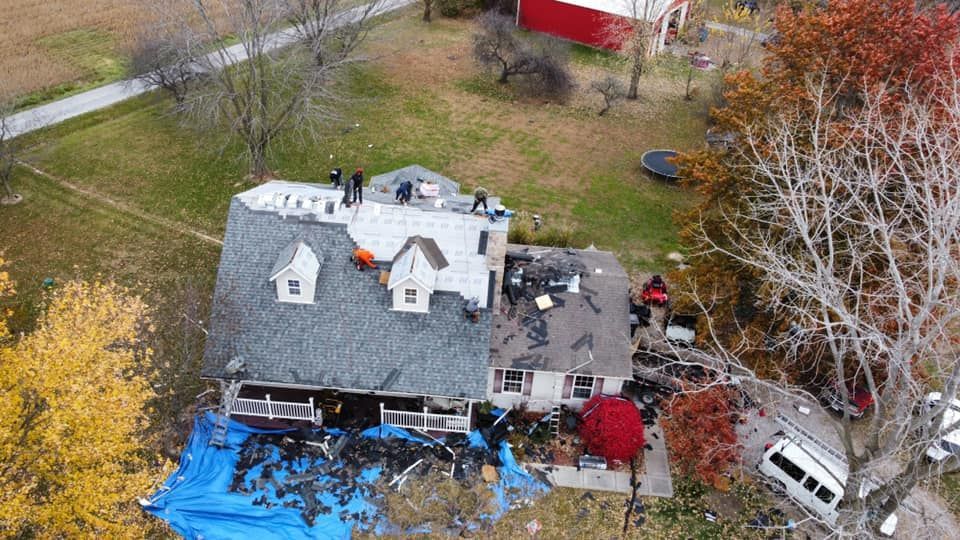 Aerial view of a house with roof damage and workers on the roof, surrounded by trees and a lawn.