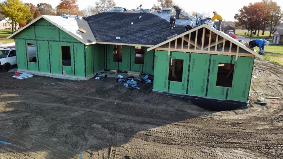 House under construction with workers on roof and ground. Green exterior walls, black roof.