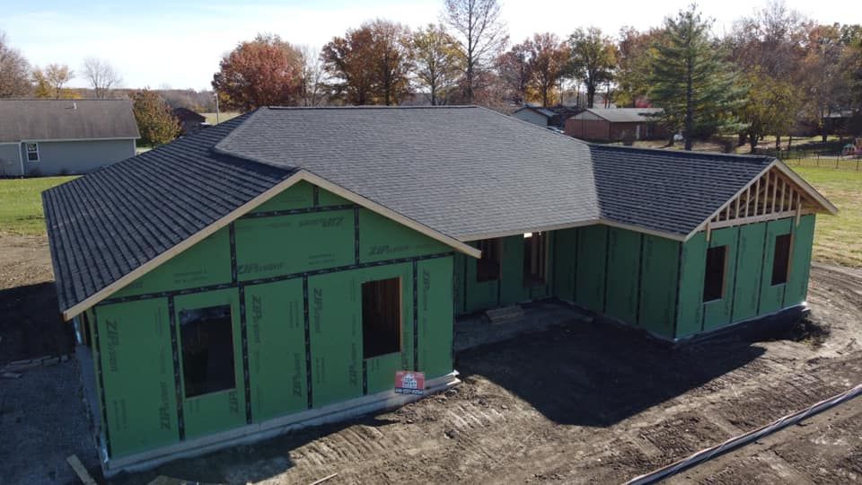 Unfinished house with green sheathing, dark roof, and windows, surrounded by dirt and grass.