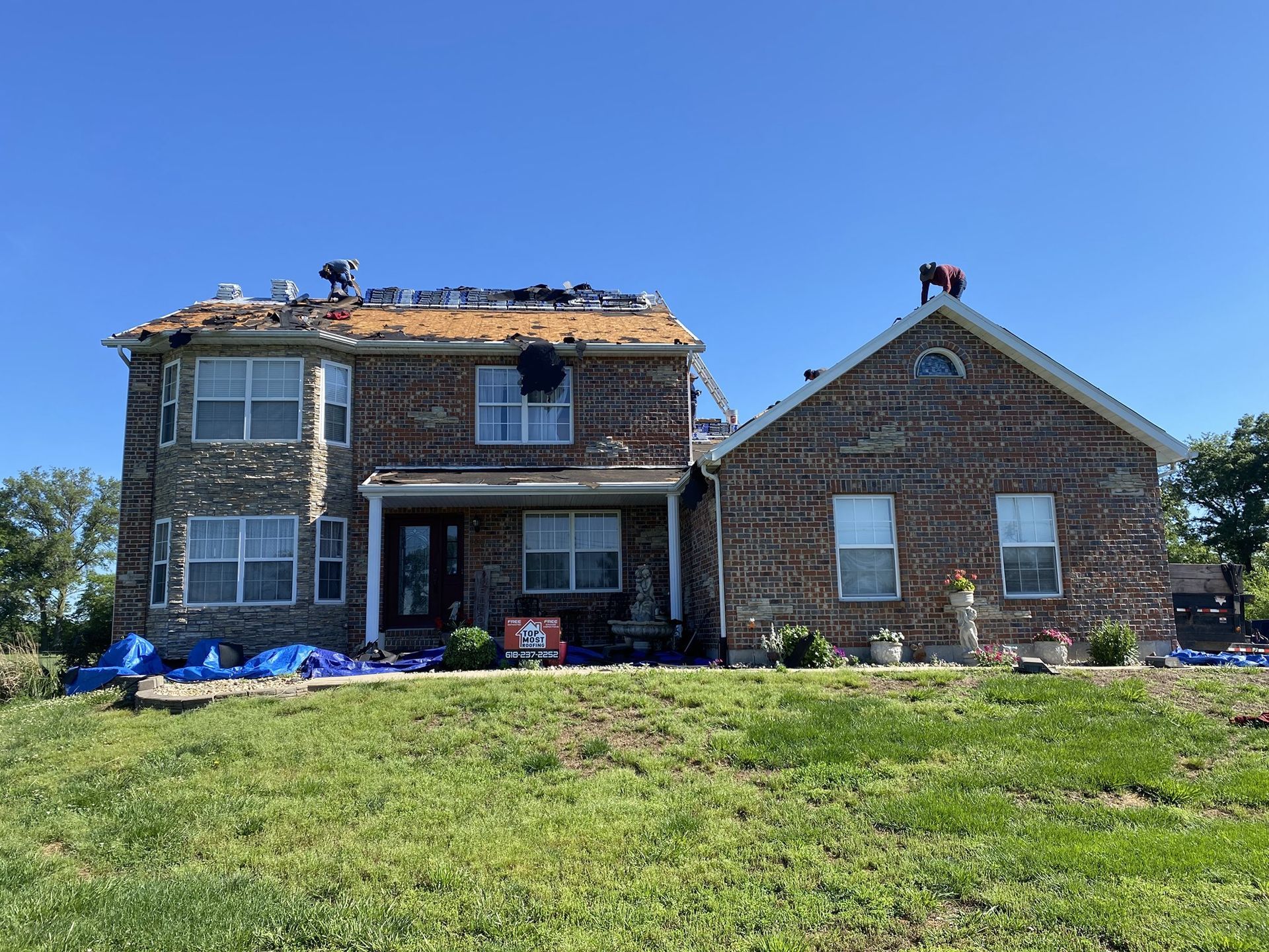 Damaged brick house; roof torn off, covered with tarps. Two figures on roof, bright blue sky.