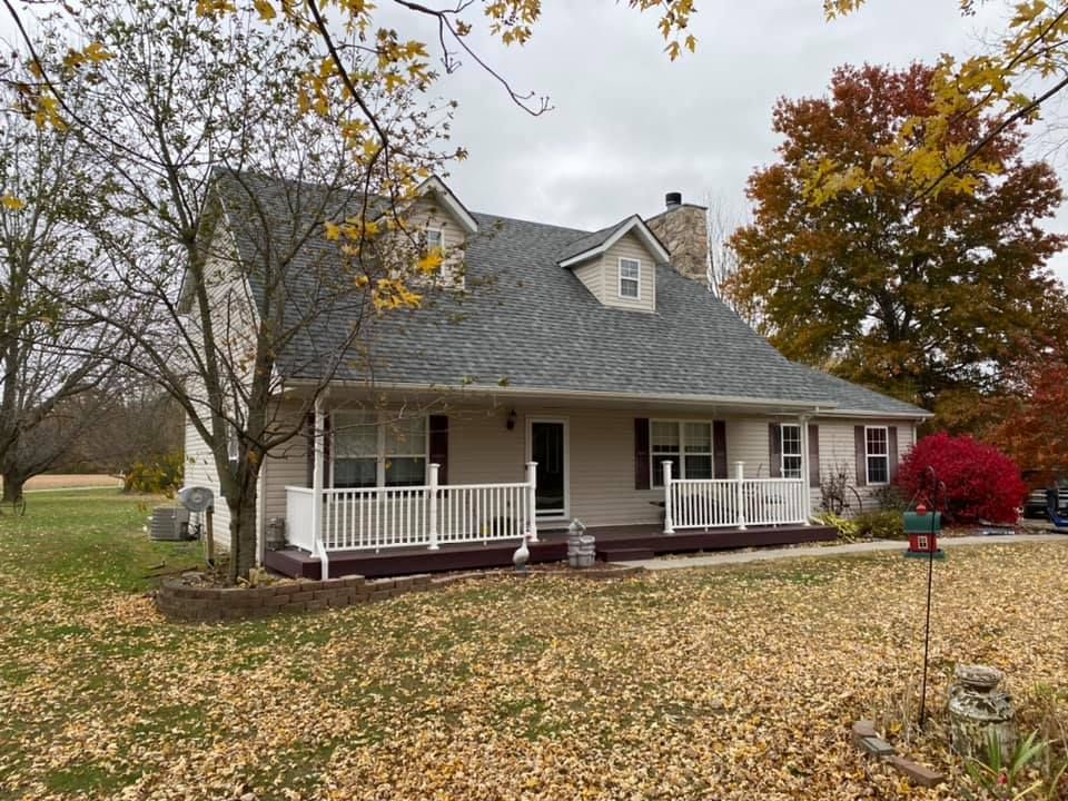 Two-story house with porch, gray roof, and chimney. Fall foliage surrounds the home on a cloudy day.