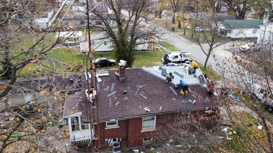 Roofers repairing a damaged roof on a brick house. Debris and vehicles are visible nearby.