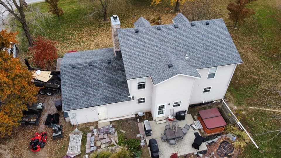 Overhead view of a two-story house with gray roof, a patio with furniture, and vehicles in a yard.