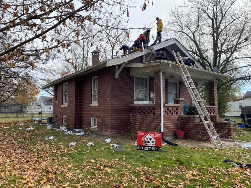 Workers on a brick house roof during a roof repair. A ladder leans against the porch. Fall foliage.