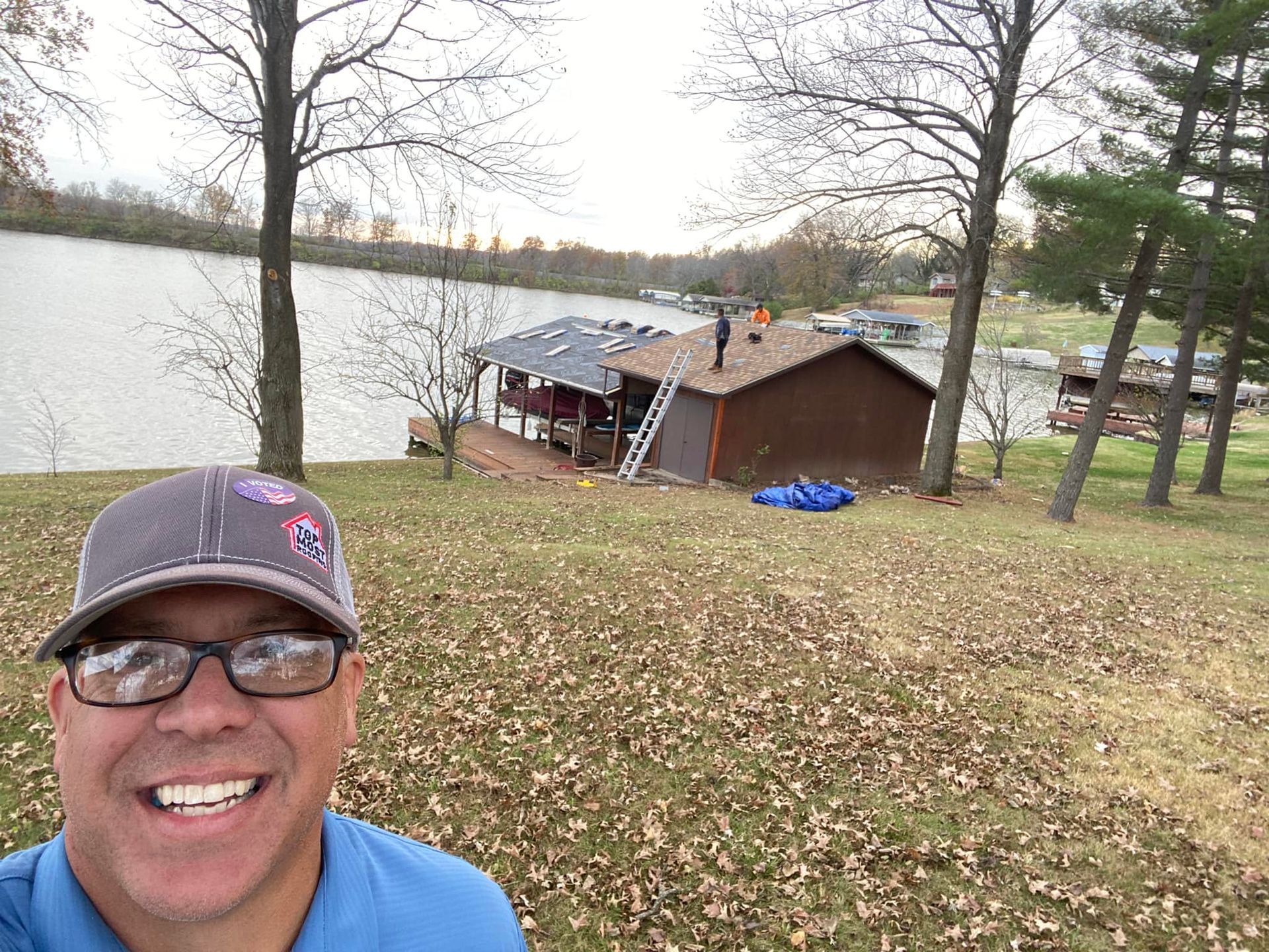 Man smiling with a lake and roofing work in the background. Brown building and trees.